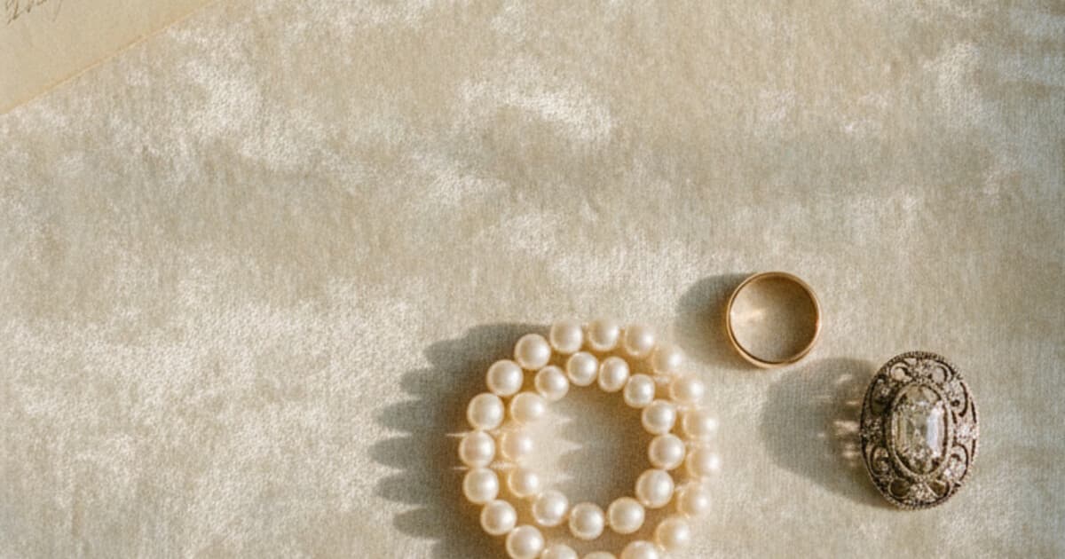 Overhead flat-lay on aged cream velvet of a mixed jewelry collection representing a family inheritance: gold ring, pearl necklace, vintage diamond brooch, and a folded handwritten letter with warm natural window light