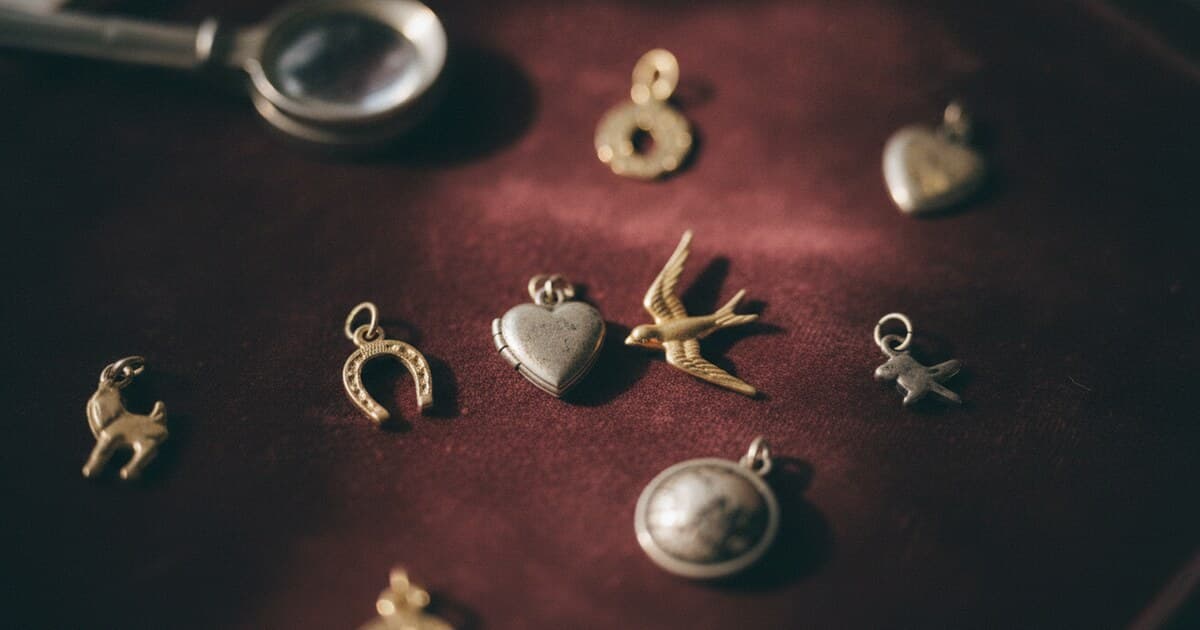 Collection of vintage gold and silver charms arranged on aged burgundy velvet, including a horseshoe, heart locket, and swallow charm, with a jeweler's loupe nearby